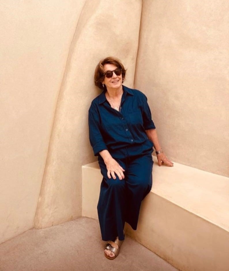 #ParaTodosVerem: Mulher sentada em um banco de concreto claro, encostada em uma parede com formas curvas na mesma tonalidade bege. Ela usa óculos escuros, camisa e calça de cor azul-escura, além de sandálias claras. Está sorrindo levemente e apoiando uma das mãos no banco. O ambiente é minimalista, com tons neutros e iluminação suave, destacando as texturas do espaço arquitetônico.